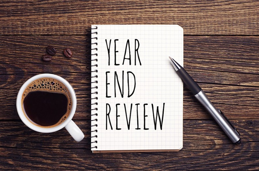 Notebook that has "Year End Review" written on it. Pen is leaning on the notebook and a full coffee mug besides it. On a dark wood background